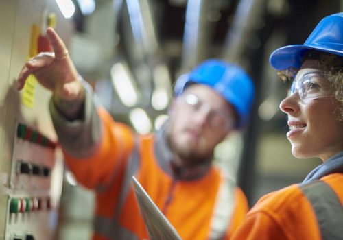 two industrial service engineers conduct a safety check of a control panel and boiler room at a power station . They are both wearing safety equipment and are looking at a control panel .